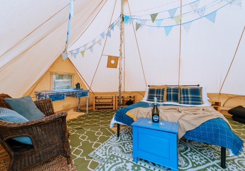 The inside of a cotton canvas bell tent, a double bed is in the centre of the tent dressed with bed linen, up-cycled furniture is on display and woven accent chair in the corner. Green pattern rugs are used as the flooring base inside the tent,
Layers of pale blue and light green bunting hang between tent poles.