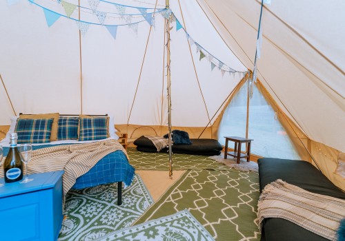 A colourful interior of the bell tent with 4 patterned rugs underfoot. Cotton Canvas tent sides meet the high emperor ceiling. Pale blue bunting and striped blankets placed inside the tent. Four floor mattresses, two are stacked together on each side of the tent. 
A dressed double bed with additional blankets and pillows.
