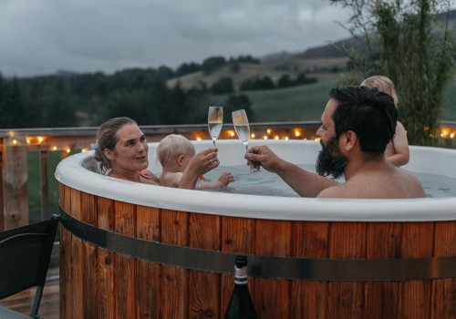 A young family of 4 are sitting in the large wood fired hot tub at sunset with fairy lights around the wooden veranda whilst Mum and Dad cheers their champagne flutes and children splash in the hot tub behind them. Their views are looking over the Cairngorm national park from Glenshee Glamping