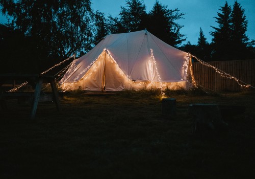 Night time photo of the cream canvas bell tent, lit up at night with warm fairy lights along the guy ropes and outside the A-Frame tent. Silhouetted pine trees sit behind the bell tent with a light blue evening sky.