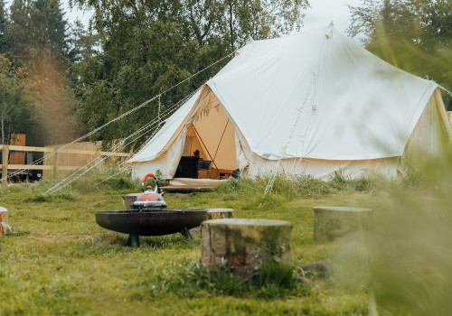Cream Canvas bell tent accommodation pitched on green grass and seen through blurred foliage. The rolled back tent curtains opens to show furniture in the distance. An iron firepit with a red steam kettle sit in the middle of the grassed area surrounded by natural tree logs for seating