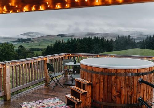 A fully wooden hot tub with its own raised veranda, lit up with cosy lights. Light steam coming off the hot water and low level clouds hitting the rolling hills in the background make for a moody, yet cosy atmosphere. A metal frame table and chairs for two sit in the corner edge of the veranda with a bottle of champagne and two glasses.