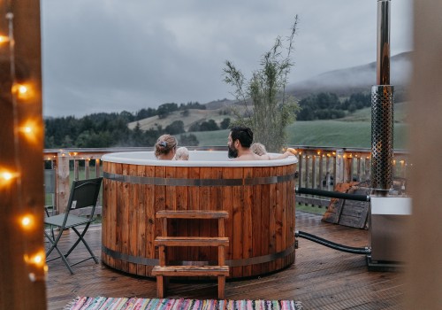 A long focus image of the cosy large wood fired hot tub, fully wooden complementing steps to enter the hot tub. A family are sitting in the water in the hot tub, surrounded by warm fairy lights for a secluded atmosphere whilst looking at their view of the rolling hills and low clouds for a moody scene at Glenshee Glamping