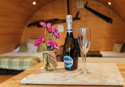 A picture of Welcome champagne, two glassed and a jar of freshly picked flowers from Glenshee Glamping fields sit on the kitchenette bench as you enter the Mulberry accommodation cabin