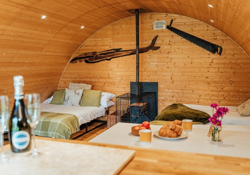 A cosy rounded roof cabin, fully wooden throughout, with a double bed placed to the far left and two single beds to the right hand side. A family sized breakfast bar and benches are dressed with fresh pastries, fruit and coffee. A log burning stove beside the beds and a bottle of champagne is sitting on the kitchenette Work top as you enter the cabin. Olive green textiles and natural furs decorate the glamping accommodation